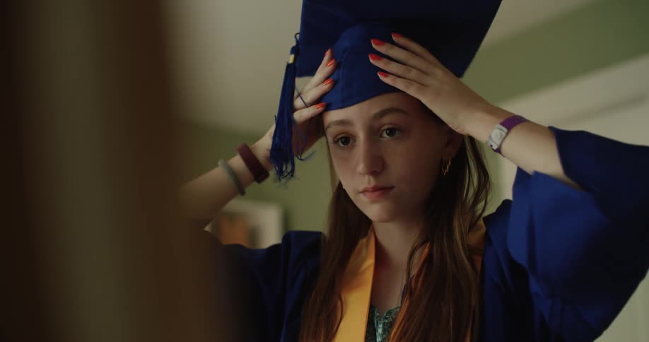 A teenage girl puts on a graduation cap and looks in the mirror