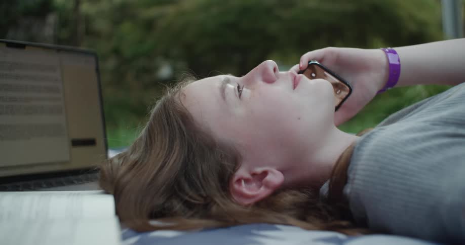 Close up of a teenage girl laying on a blanket outside while talking on her cellphone next to her laptop