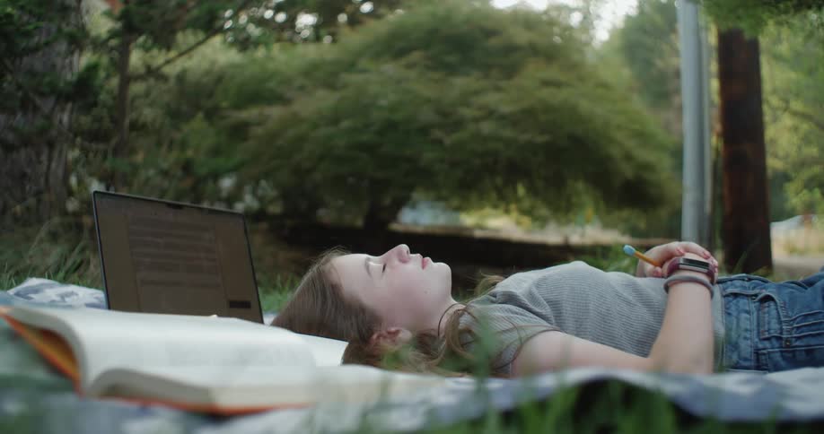 Student lies on blanket with laptop and textbook and rests on her back