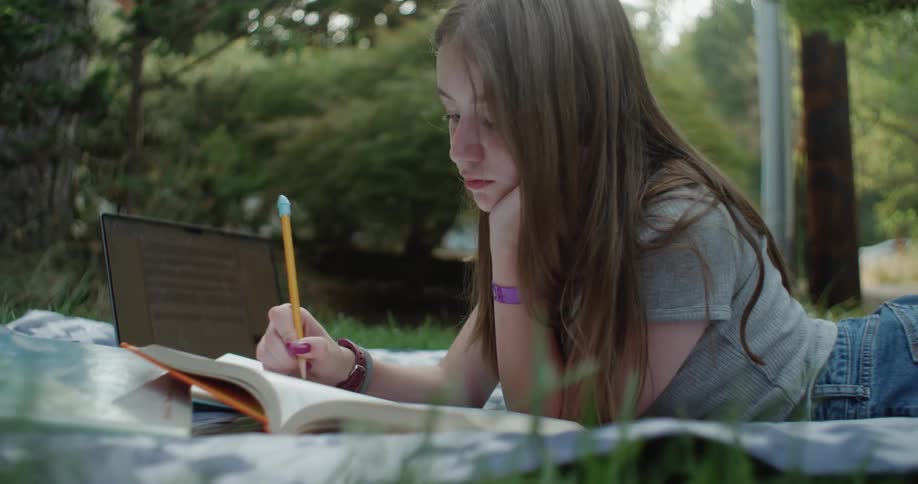 Young teenage girl laying on a blanket outside flipping through a school book writing with a pencil near a laptop