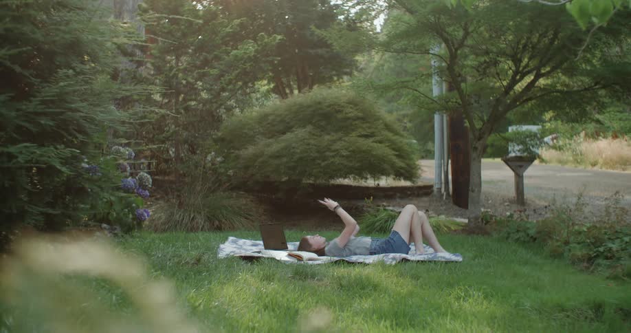 A teenage girl is lying on a blanket in her yard with a laptop computer as she holds her phone in the air