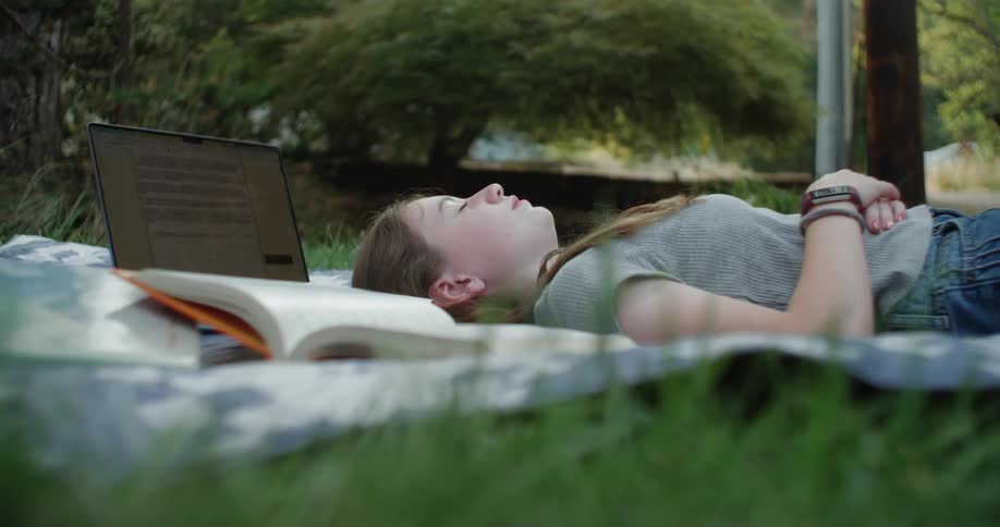 Teenage student naps on blanket on lawn next to laptop and textbook