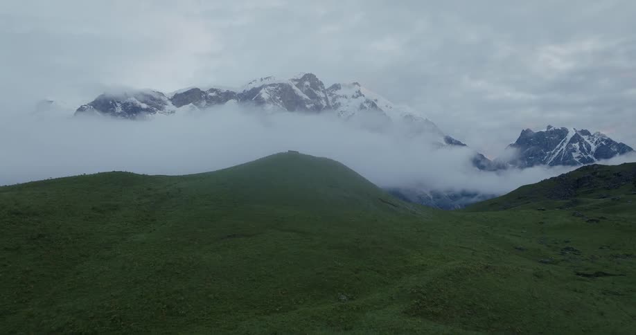 Aerial view of green hills next to a mountain range covered in clouds