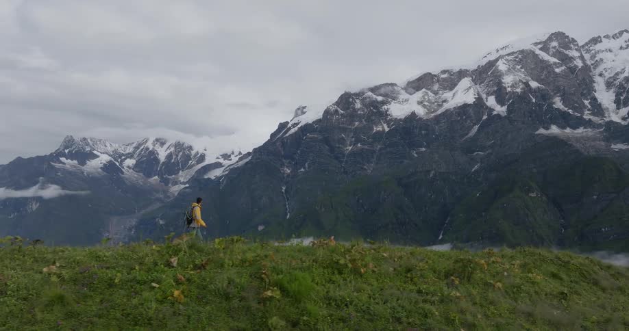 A man hiking on a grass covered mountain peak with large mountain ranges covered in snow in the distance