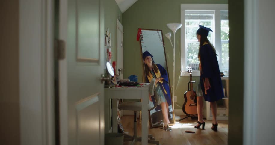 Teenage girl wearing a graduation outfit looking at herself in a mirror in her room twirling around