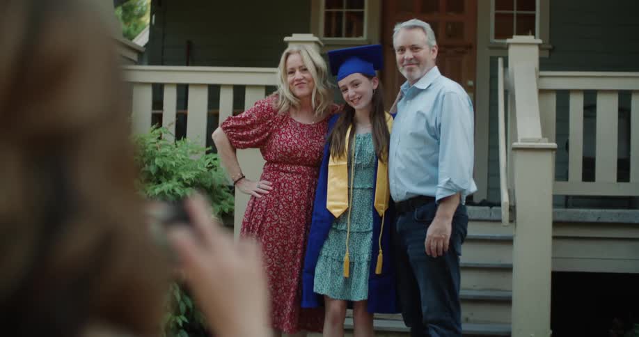 Woman takes picture of parents posing in front of house with daughter wearing a graduate robe