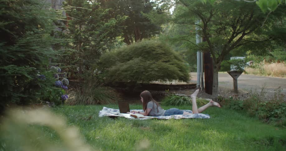 Young girl doing homework on a laptop over a blanket in the backyard
