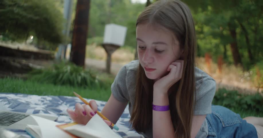 A teenage girl is lying on a blanket in her yard reading a book as a car passes by on the street