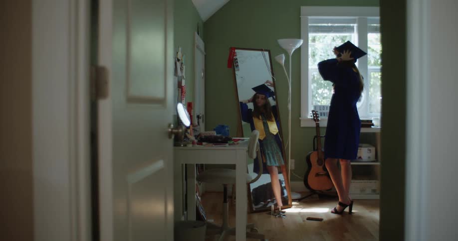 A teenage girl dressed in graduation gown looking at herself in the mirror