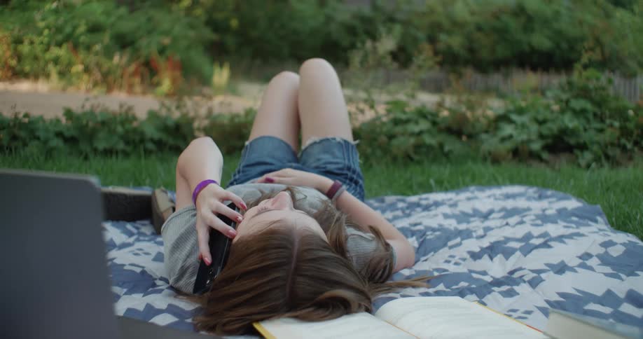 A young woman reading a book while resting on a blanket outdoors makes a call on her smartphone
