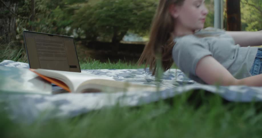 A teenage girl texting on a blanket then turning over and looking at her books and laptop