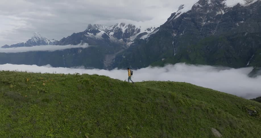 Drone view of a male hiker walking on a grassy mountain trail as fog settles in the valley below snow covered mountain peaks