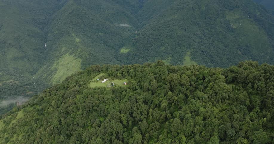 Drone view of a forest covered mountainside with rural buildings in a cut out area with mountains on the landscape