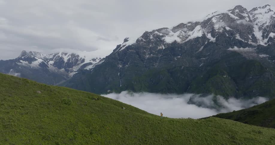 Aerial of female hiker walking up green mountain slope with snowy mountain range in the background
