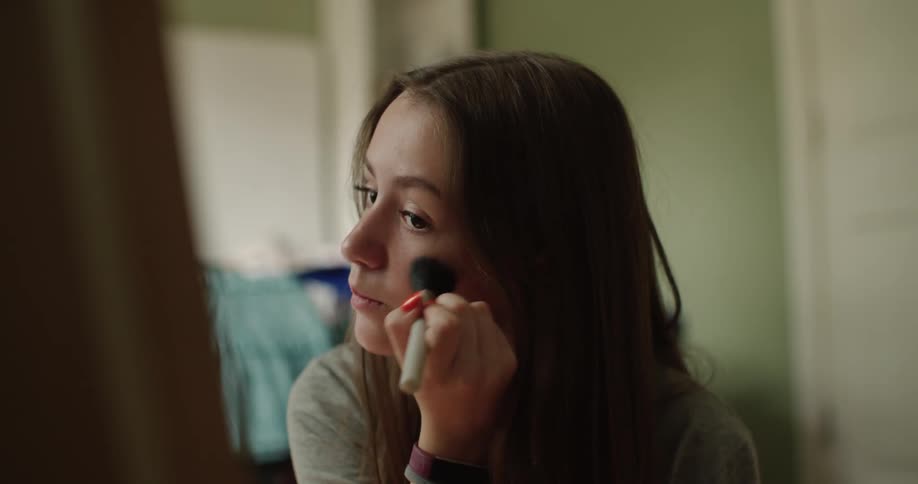 A teenage girl is sitting in front of a mirror putting blush on her cheeks with a makeup brush