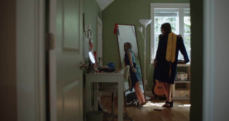 A teenage girl trying on graduation cap and gown in her room in front of the mirror