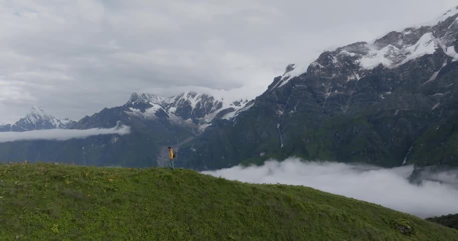 A man is walking on a grass covered mountain with snow covered mountains in the distance under a cloudy sky