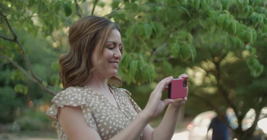Woman standing under a tree holding her phone up to take a picture as she smiles and tilts her head