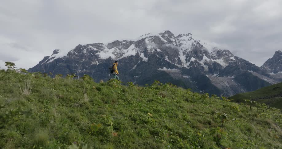 A man with a camping backpack walking on top of a grassy hill and behind him is a huge mountain with its peak covered in snow