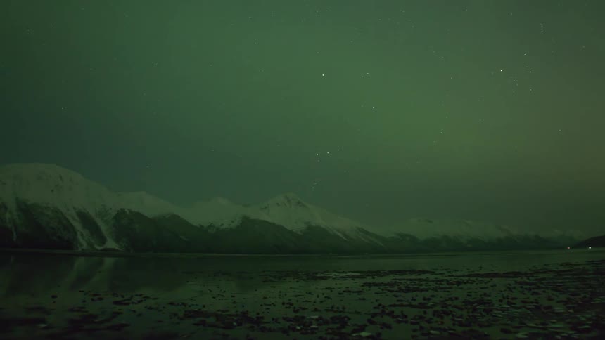 Motor vehicle lights move along highway as a plane flies over lake near snowy mountains at night