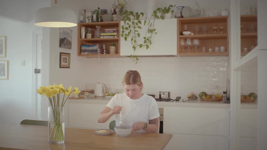 A woman sitting in her dining in her kitchen preparing noodles with a plate and spoon