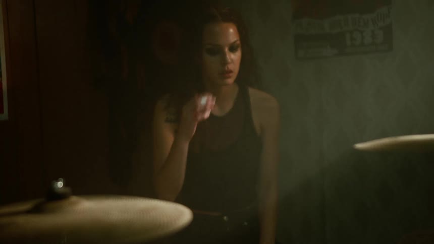 Woman with brown curly hair and black top sits down at drum kit