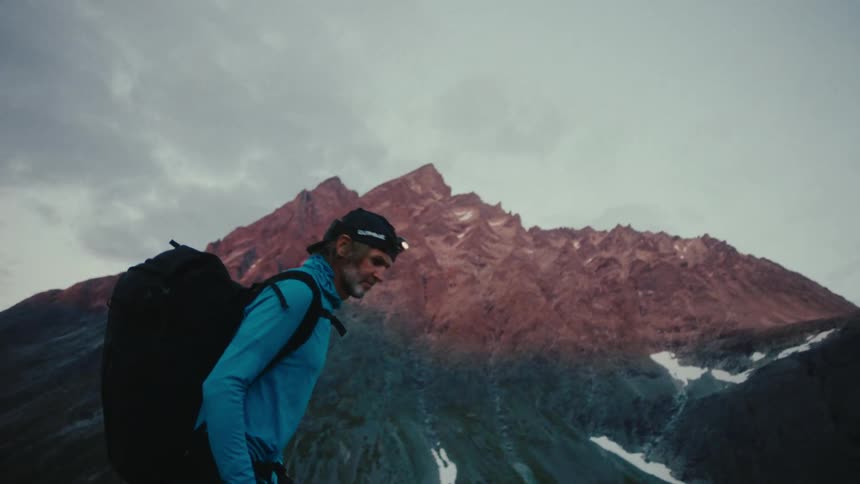An adult man walking near a mountain and is wearing camping clothes and equipment