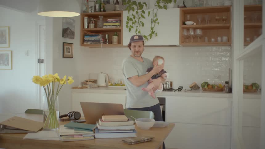 A father feeding his child with a bottle while using his laptop in the kitchen at home