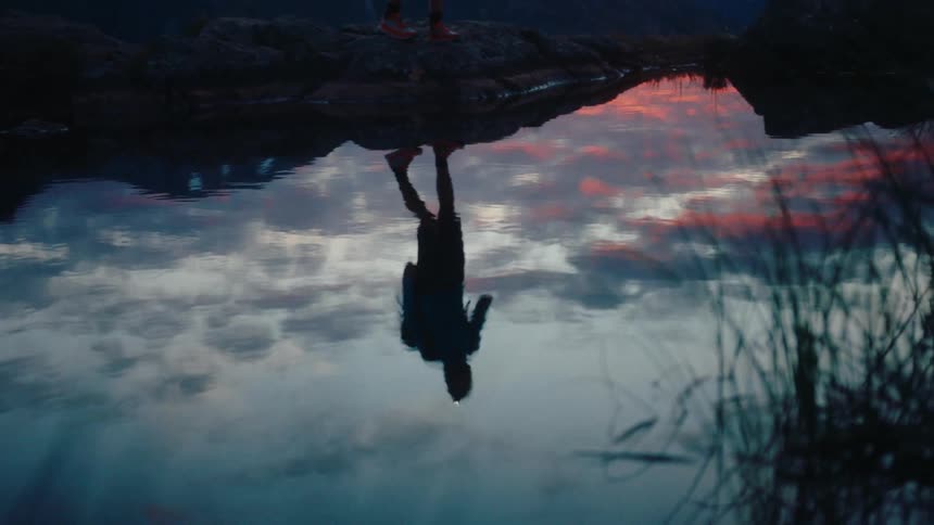 A person walking along the shore of a lake with their reflection in the water at dawn in the mountains