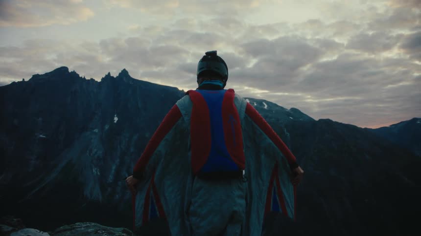 A man opens the wings of a wingsuit on top of a mountain at evening