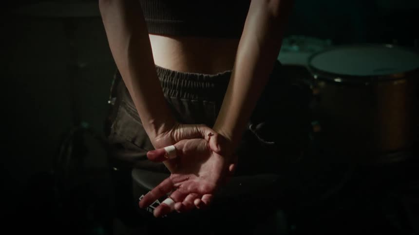 A person sitting before a drum set in a dark room stretches their arms behind their back