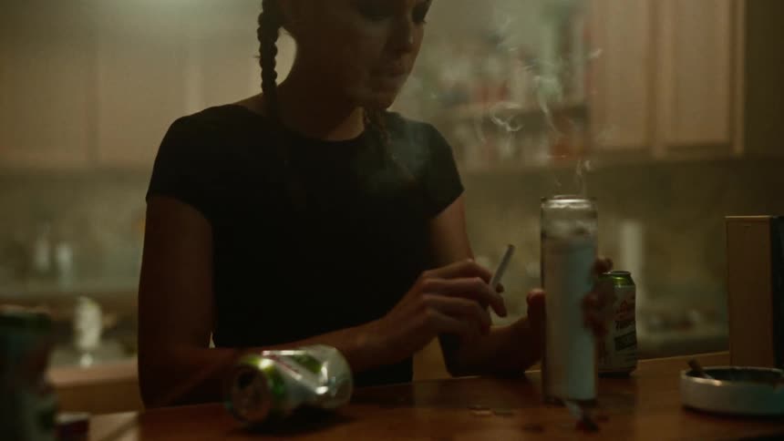 A woman is standing at the kitchen counter as she lights her cigarette from a candle and picks up a can of beer
