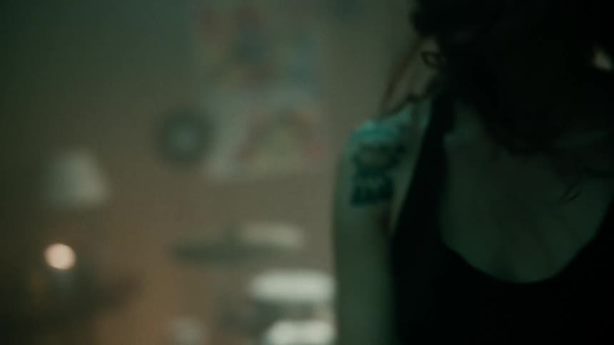 A teenage girl is walking across a room near drums and shaking her head up and down as her hair flies and she raises her arms up and down
