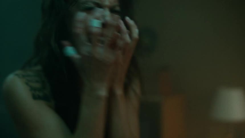 A young woman with eye makeup looking straight ahead screams and puts her hands in her hair nervously