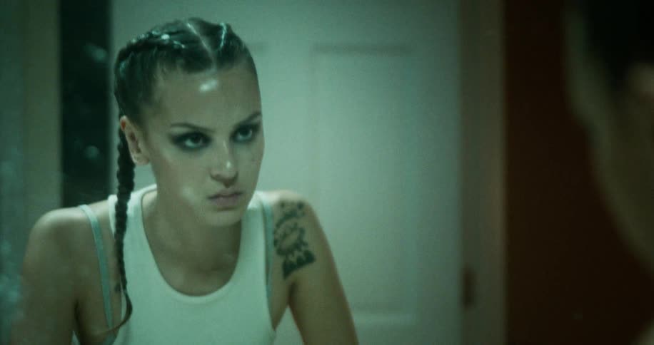 An angry looking teenage girl walks through a house to a mirror and scowls as she looks at herself