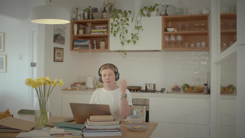 A woman is sitting and takes a headset and puts it on her head and waves to someone she is talking to on the computer on the table