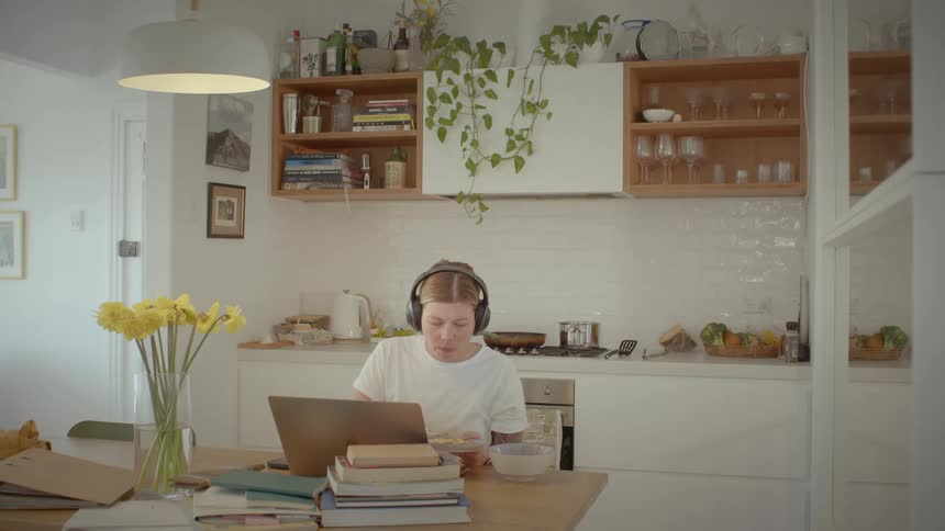 A woman sitting eating pasta and working on her laptop in the kitchen