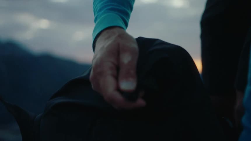 Tan gnarled hands roll up the top of a backpack and secure it on a rocky mountain ridge