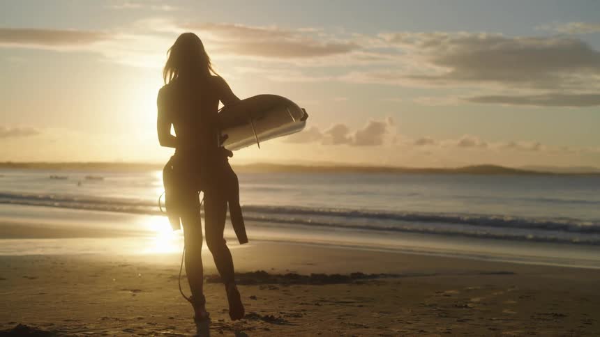 Female surfer walks toward setting sun while carrying a surfboard