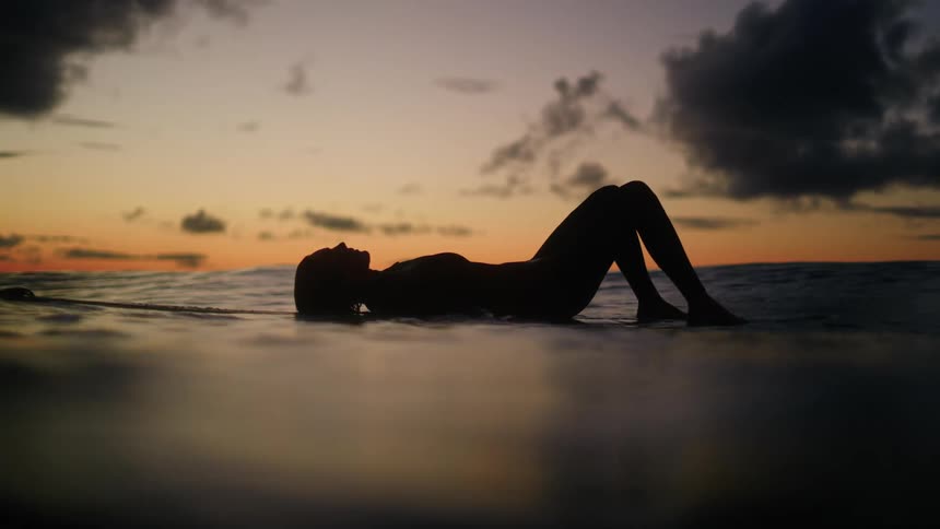 A woman in the sea at dusk lying on a surfboard on her back