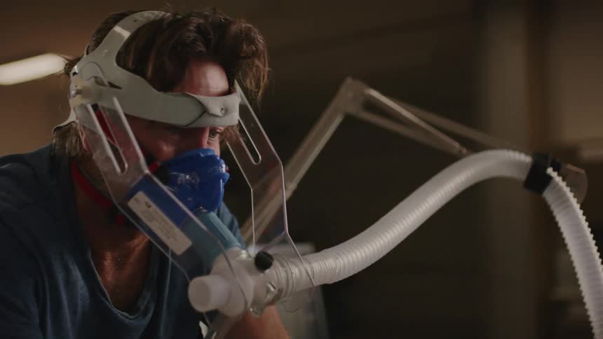 A man is pedaling a physiotherapy bike while he has his face intubated and a doctor stands beside him