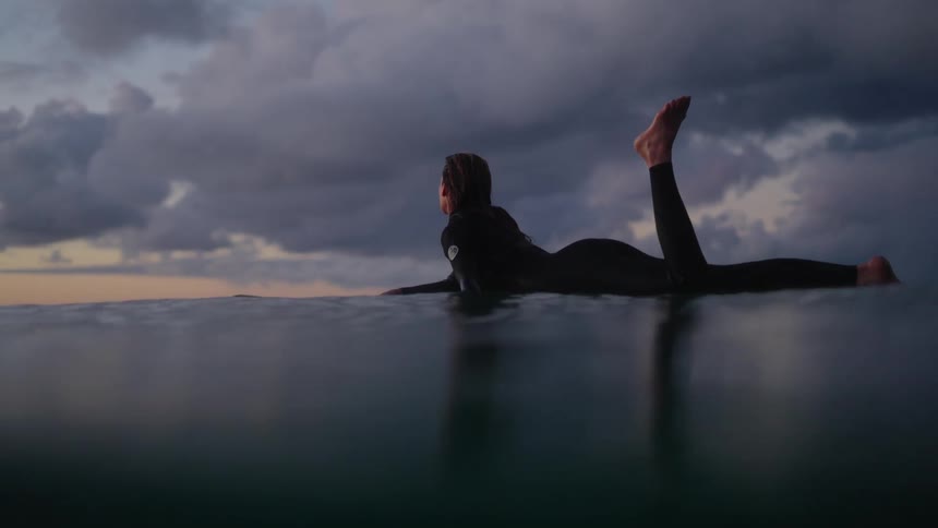 Woman floats over a surfboard in the ocean at sunset