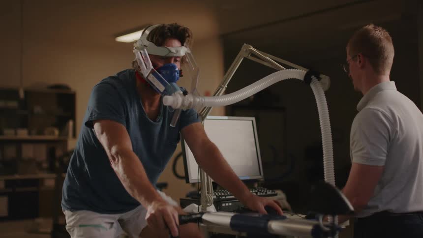A man on a medical bicycle with a breathing mask on while a medical professional stands next to him talking to him