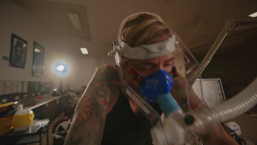 Blondek woman with tattoo and black tank top exercises on stationary bike wearing bipap mask