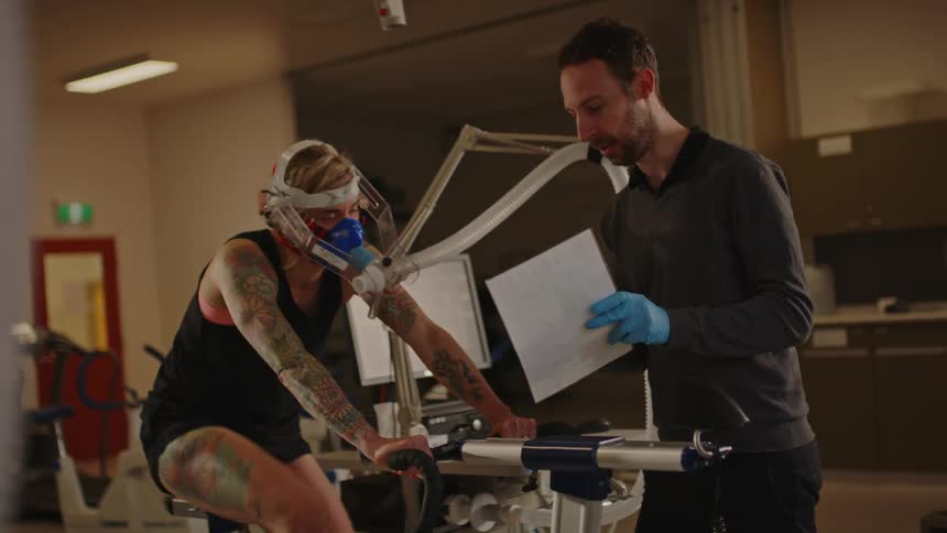 A woman with a mask on her face attached to a tube pedals on a stationary bicycle in a medical setting