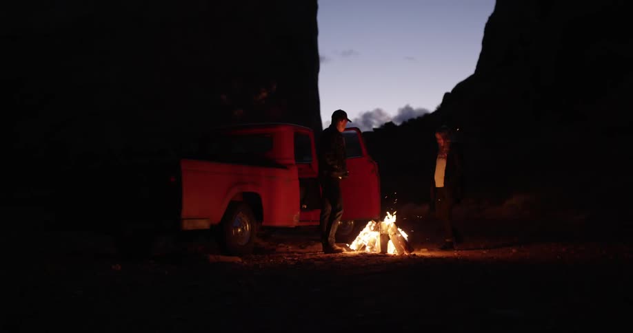 A campfire is near a truck with two people standing nearby