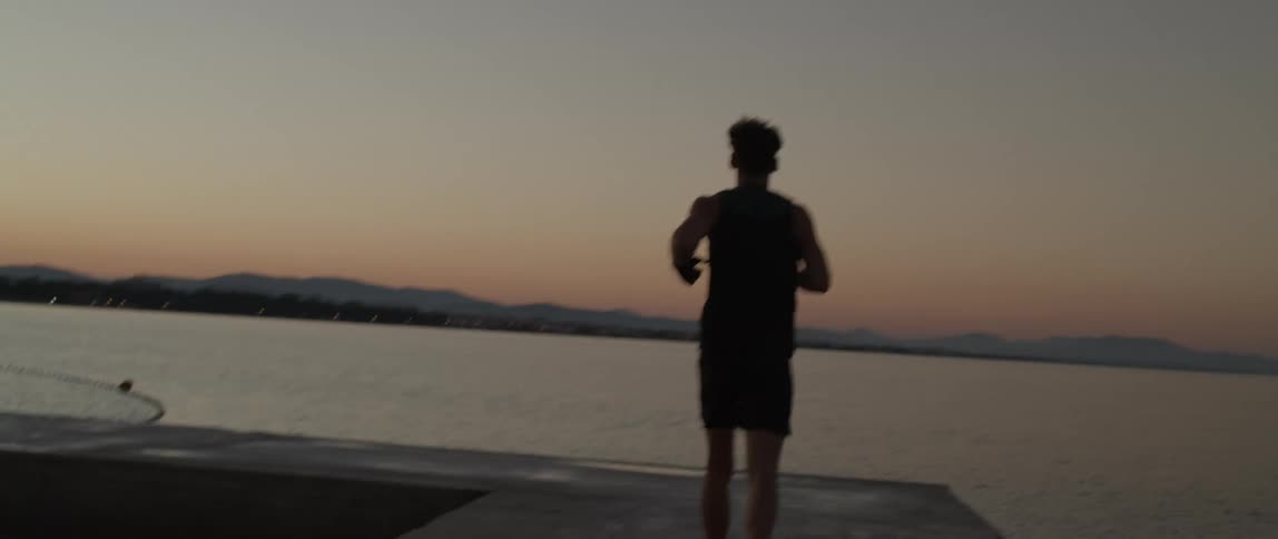 A man running with his back to the camera on a seaside dock and practicing boxing movements at dawn