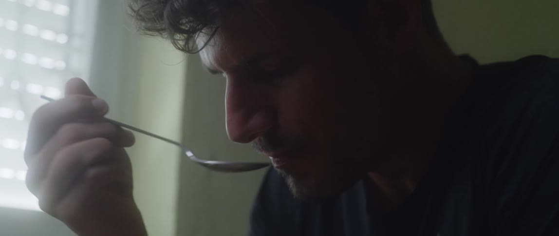 A man sits near a window while eating a bowl of cereal with a spoon