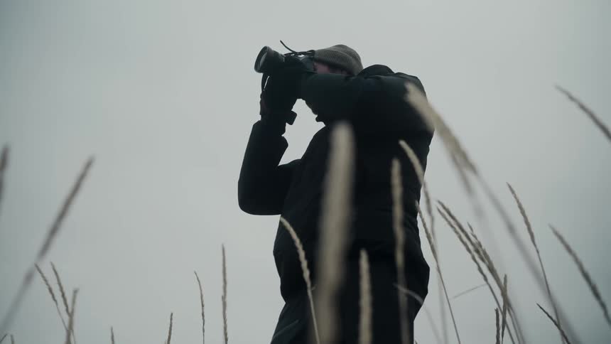 Photographer holds camera to their eyes while standing outdoors and tall grass