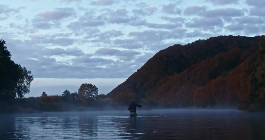 Fisherman casts his line while wading in lake near mountain in autumn on a cloudy day